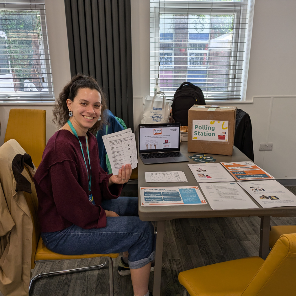 Styliana sitting at a desk covered in stacks of paper handouts while she hold us the ballot papers. 