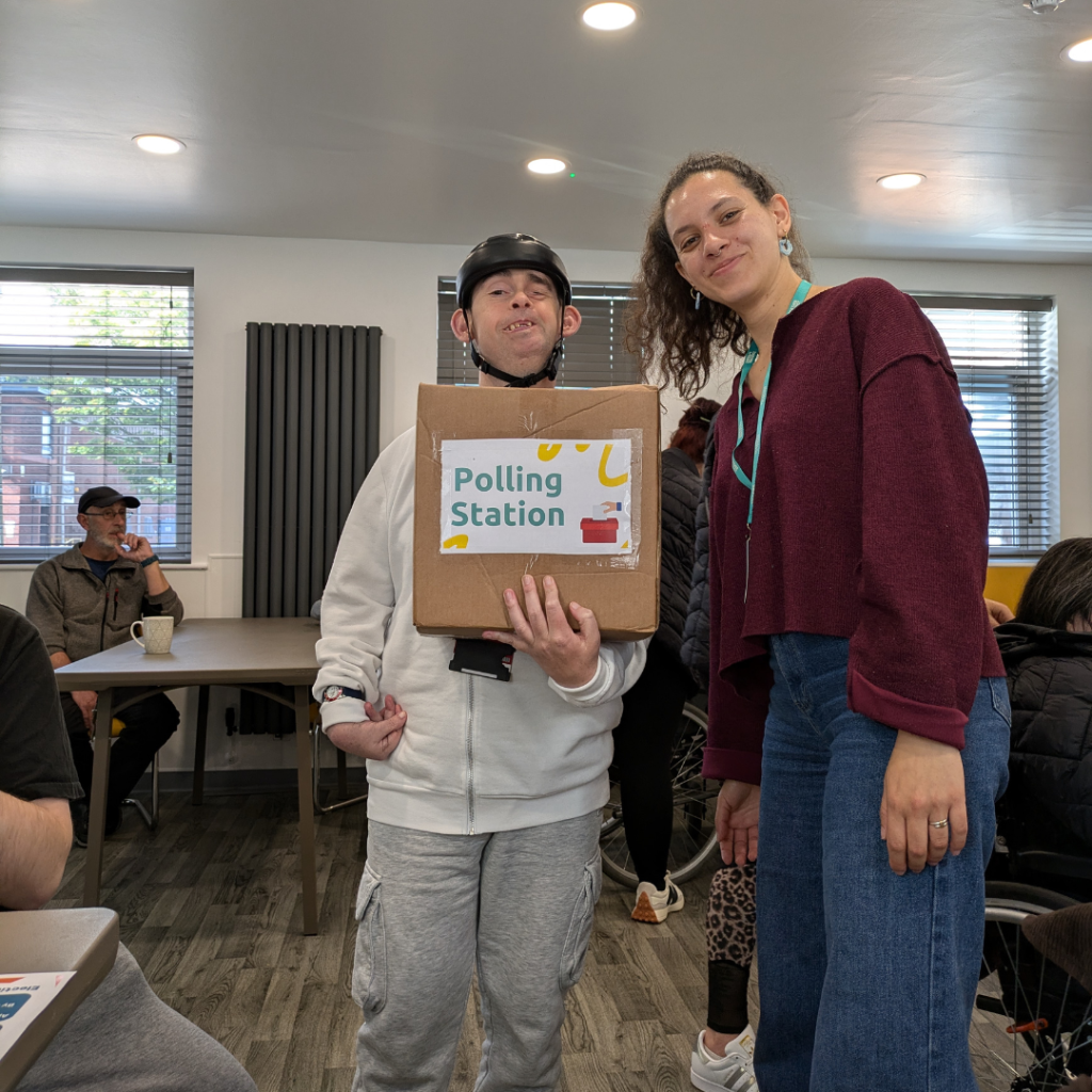 Styliana and Mike, one of the adults we support, holding a box that reads "polling station"