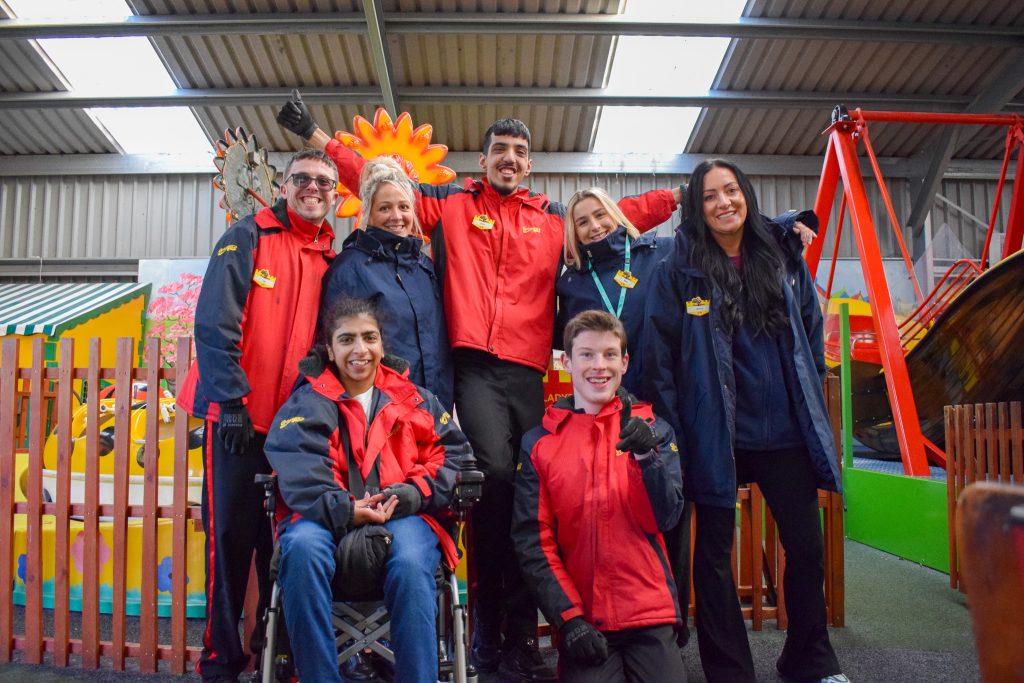 A group of 4 interns and 3 staff members smiling together in front of a colorful play area. they are wearing gulliver's world jackets.