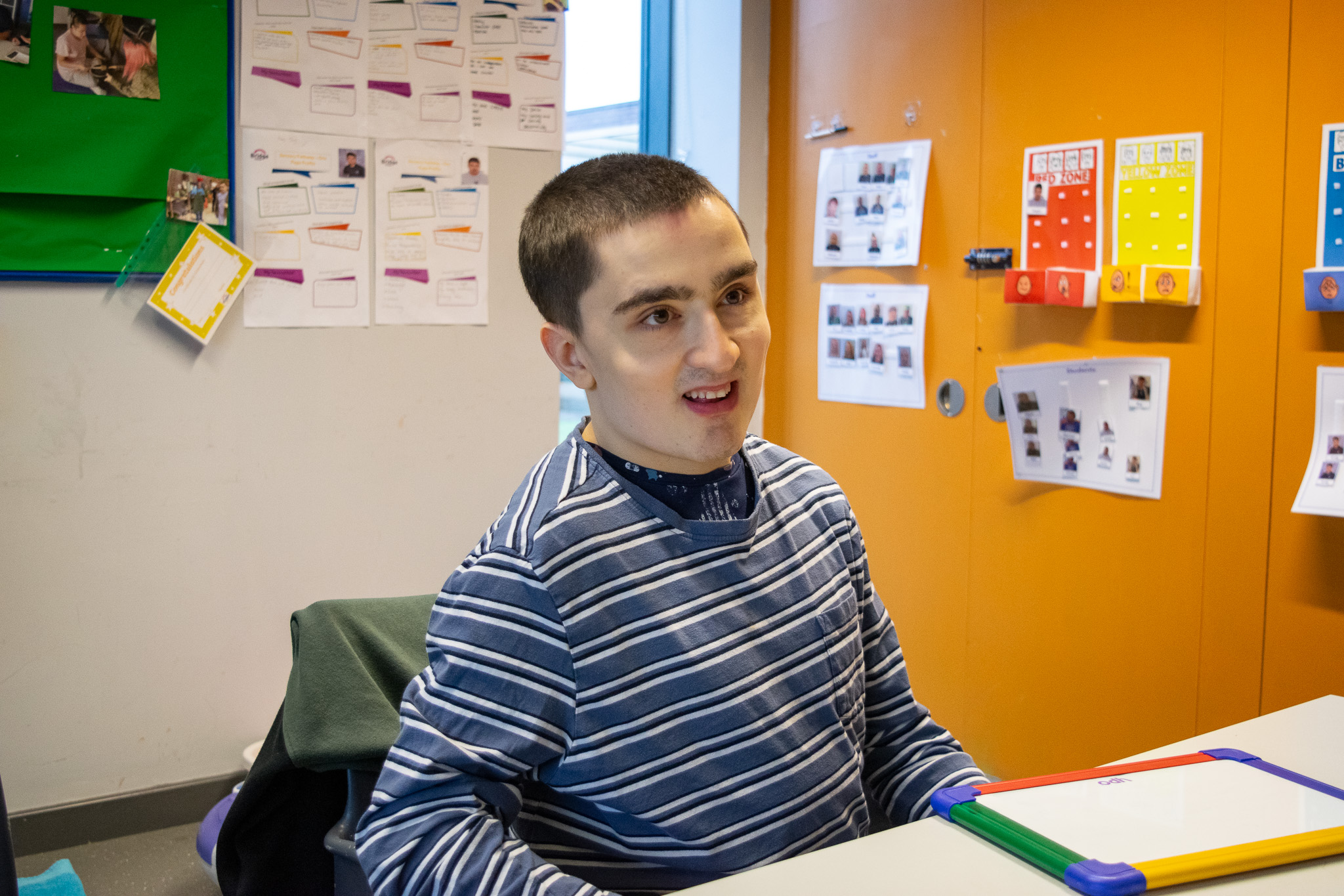 A young man is smiling as he is sat down at a desk in a classroom.