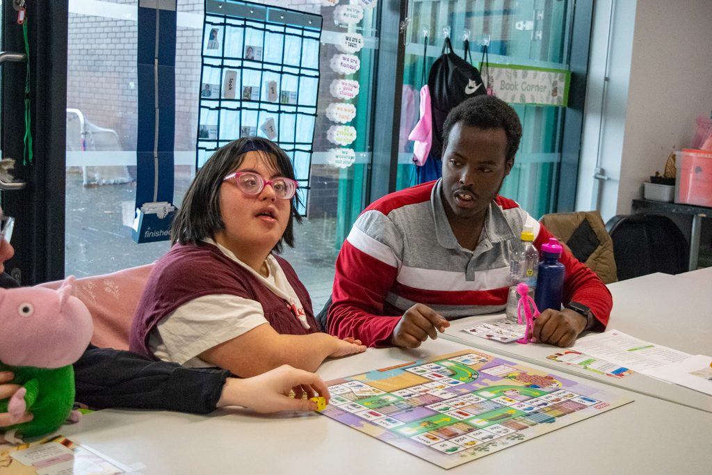 Two students sit side by site at a table in a classroom playing a board game. 