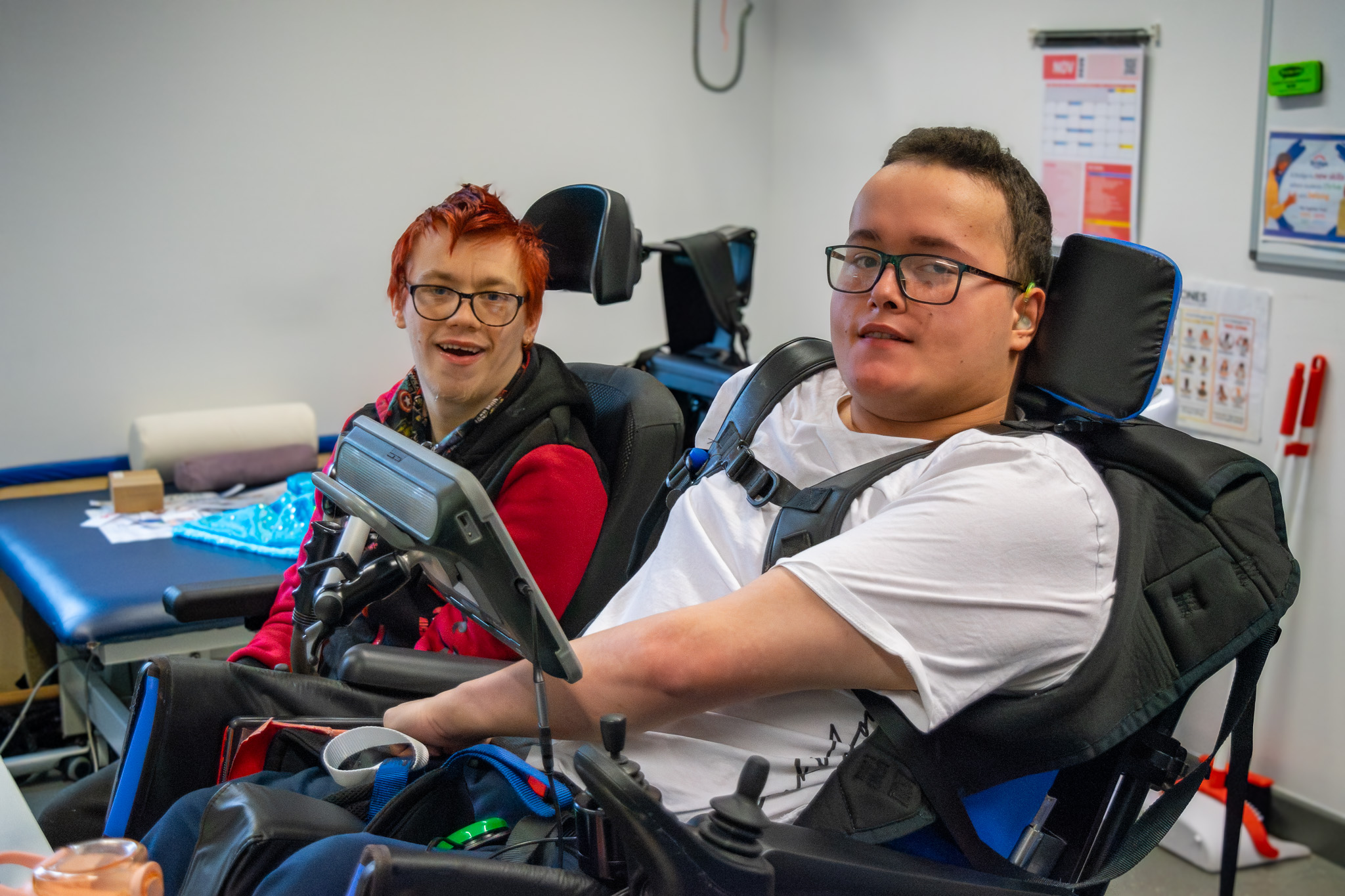 Two young men in wheelchairs in a classroom. They are looking at the camera and smiling