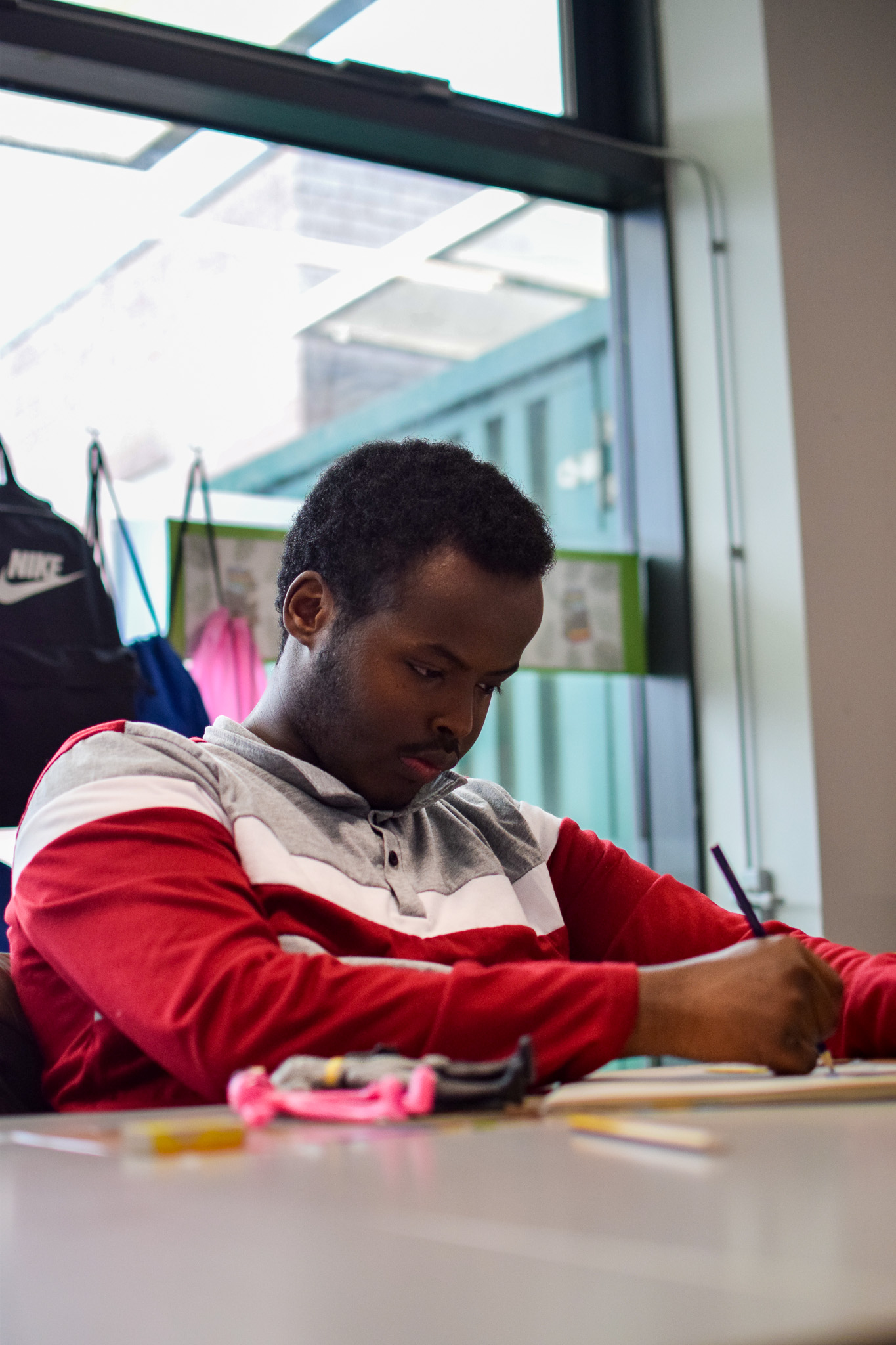 A young man is writing in his workbook, he is sat at a table in a classroom. 