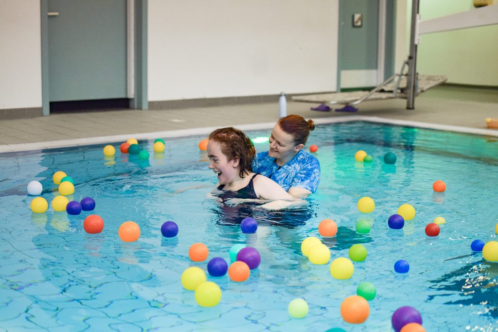 A young woman is swimming in a hydrotherapy pool with a staff member. They are playing with floating pool balls.