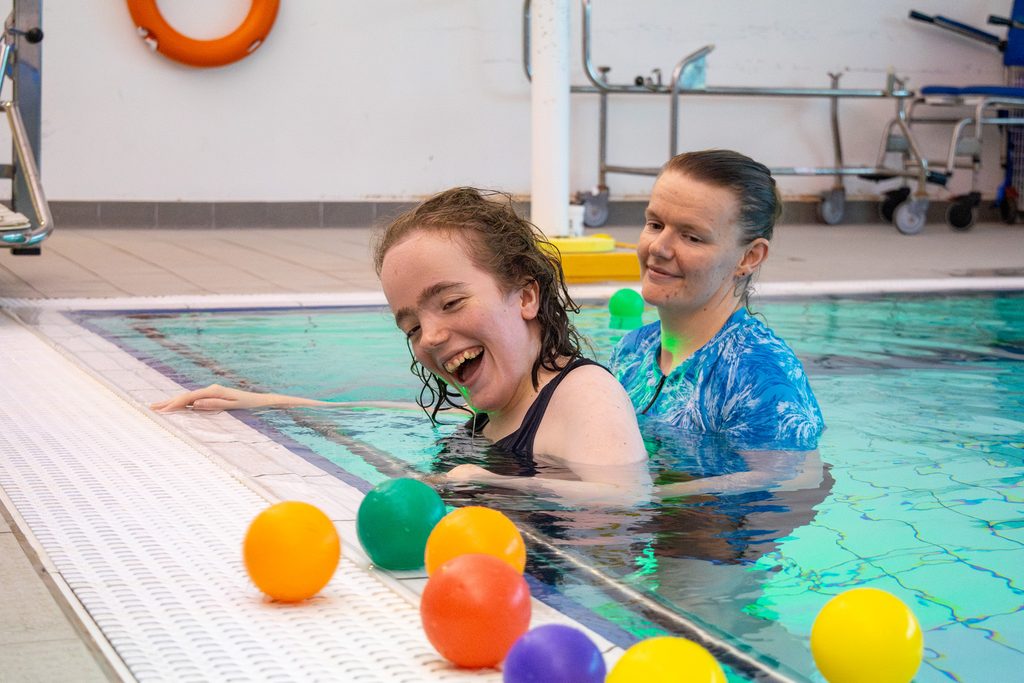 A young woman is swimming in a hydrotherapy pool being supported by a staff member. She is playing with floating pool balls. 