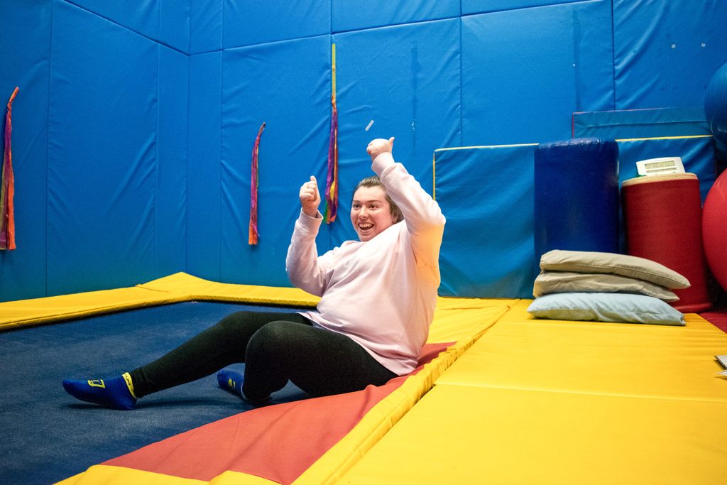 A young woman is giving 2 thumbs up after finishing a gymnastics routine in the rebound therapy room.