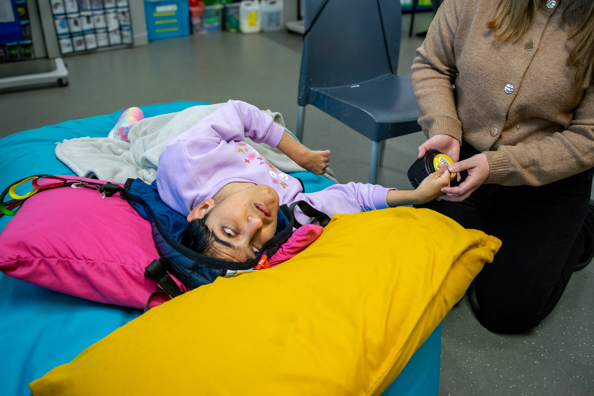 A young person is laying down on a mat while a staff member helps them press a button that emits noise. They are in a sensory storytelling class.