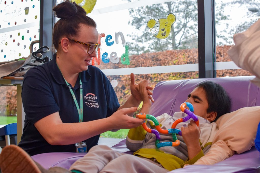 A young person is laying down on a rocking chair and playing with a sensory toy. A staff member is playing with them. 