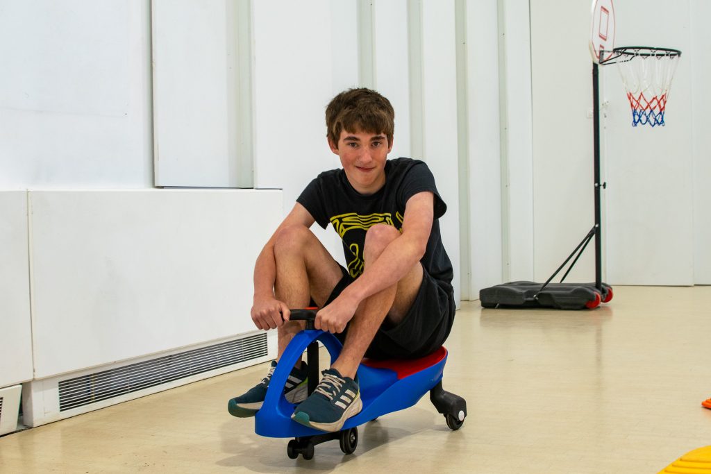 A teen boy is riding a small bike in an occupational therapy class. 