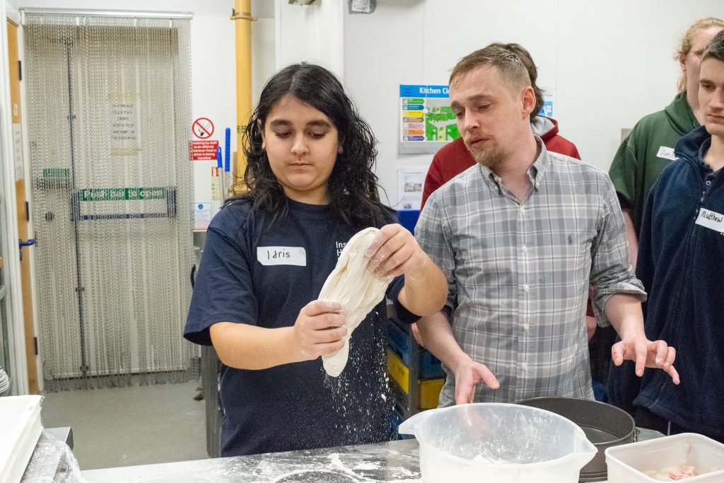 Student shaping up pizza dough in their hands while the chef is guiding him from the side. 

