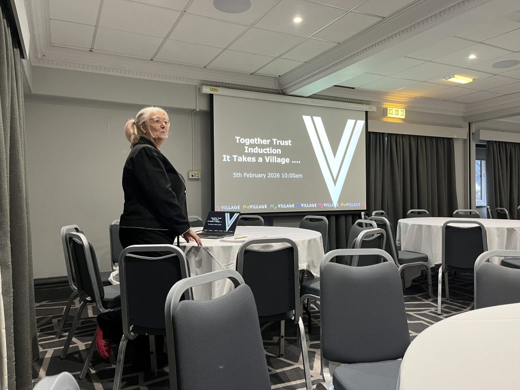 Woman dressed smartly stands in front of conference room talking to a crowd. The screen display behind her reads "together trust introduction. it takes a village"